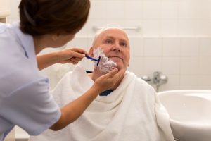 A caregiver gently assists an elderly man with shaving in a bathroom, highlighting the essential role of home care professionals in supporting seniors with personal hygiene challenges.