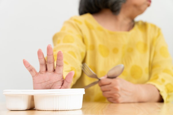 An older woman turns away and raises her hand to refuse a meal tray, highlighting the challenges caregivers face when encouraging better eating habits in elderly parents.