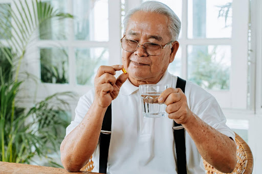 A senior man holds a pill in one hand and a glass of water in the other, smiling gently—illustrating how personal, human medication reminders can be more effective than digital app alerts.