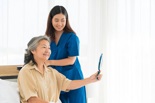 A smiling caregiver stands beside a senior woman who admires herself in a mirror, symbolizing how a gentle hand in personal care fosters dignity, confidence, and lasting trust.