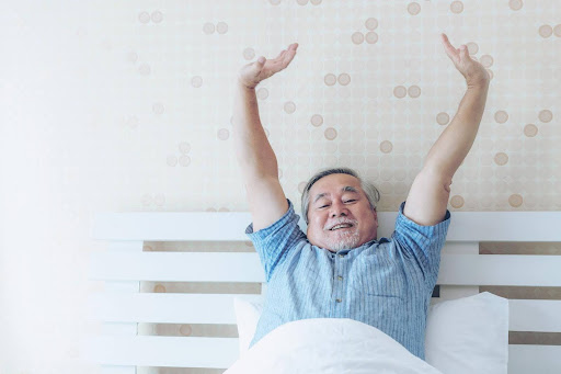 A smiling senior man stretches joyfully in bed after a good night's sleep, illustrating the positive impact of proper nutrition on sleep quality for older adults.