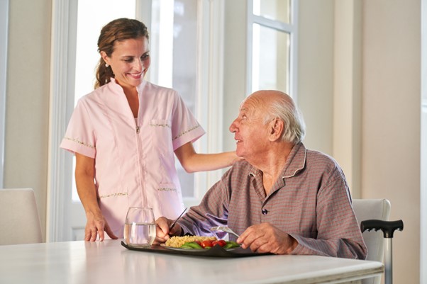 A caregiver smiles at a senior man seated at a table with a healthy meal, highlighting how meal prep services help prevent malnutrition in seniors.