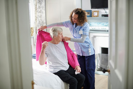 A caregiver helps an elderly woman put on a sweater in a cozy bedroom, illustrating how personal assistance supports daily tasks alongside physical therapy for overall well-being.