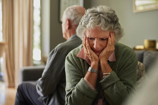 An elderly woman sits with her head in her hands, looking distressed, while an older man sits turned away—reflecting the emotional need to encourage and support aging loved ones through connection and care.