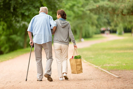 A caregiver walks beside an elderly man using a cane, carrying a grocery bag—illustrating the often-overlooked everyday tasks that in-home caregivers manage to support senior independence.