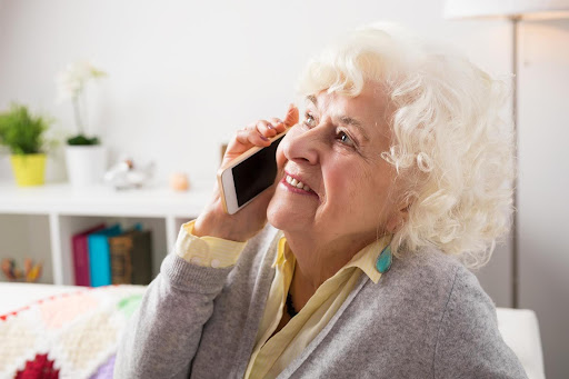 A joyful elderly woman smiles while talking on a smartphone, representing how compassionate check-in calls help bridge emotional gaps when family can’t always be there.