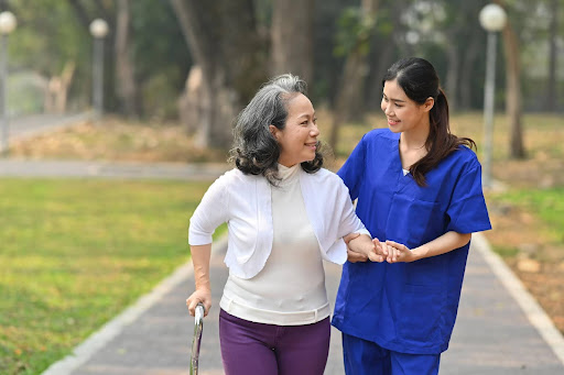 A cheerful caregiver walks hand in hand with a smiling elderly woman in a park, capturing the warmth, compassion, and dedication Filipino caregivers are known for globally.