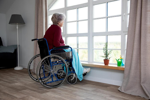 An elderly woman in a wheelchair gazes out the window of her home, alone and thoughtful—capturing the quiet guilt families may feel when living out-of-state and unable to visit often.