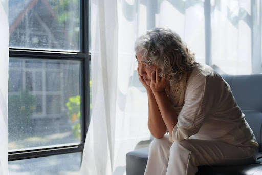 An elderly woman sits alone by a window looking out thoughtfully, symbolizing the need to explore every care option for seniors who can no longer live independently.