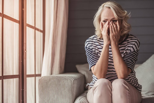 Elderly woman sitting on a couch with her hands covering her face, appearing emotional and grieving, symbolizing the loneliness some seniors feel after losing a husband.