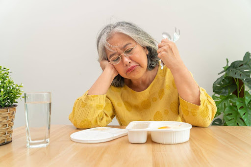 A senior woman in a yellow shirt looks upset while sitting at a table with an untouched meal in front of her, holding a fork and spoon. A glass of water is beside her, highlighting a potential issue with eating or appetite loss.