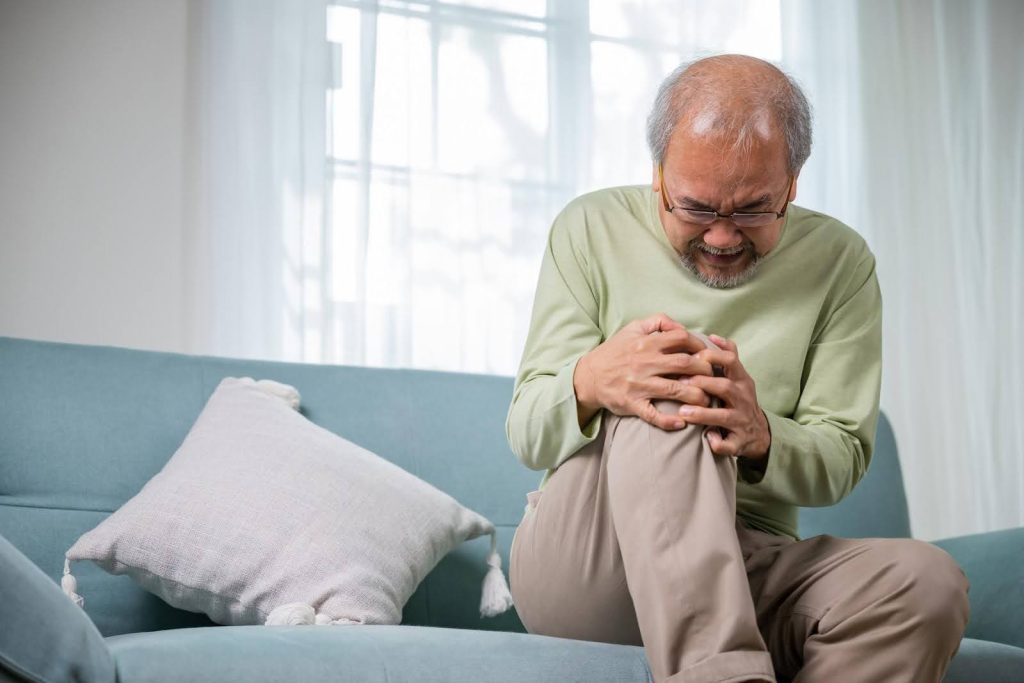 Elderly man grimacing in pain while holding his knee, illustrating the daily physical challenges seniors with arthritis face—such as difficulty buttoning a shirt or performing routine tasks.