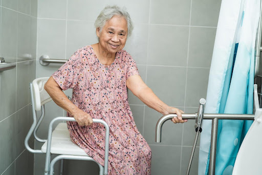Elderly woman smiling in a safety-equipped bathroom, sitting on a bath chair—showcasing personal care approaches that make bathing more comfortable and dignified for seniors.