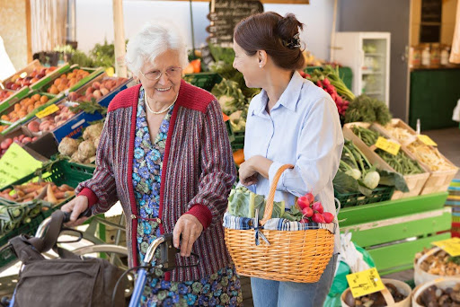 A caregiver helps an elderly woman using a walker during an outdoor errand at a local market, both smiling as they select fresh produce—highlighting compassionate errand support for seniors.