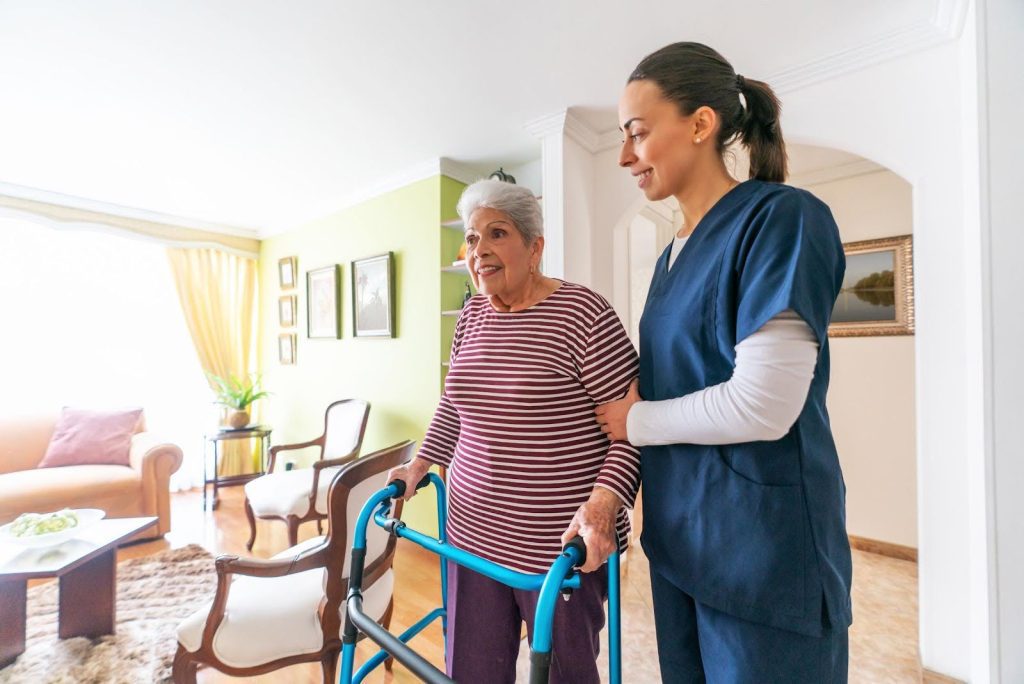 A caregiver in blue scrubs supports an elderly woman using a walker inside a bright, warmly decorated home, representing the personalized support often found in assisted living and home care environments.