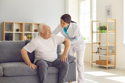 A caregiver assists an elderly man from the couch in a bright, tidy living room—illustrating the importance of a safe home setup for seniors with memory loss.