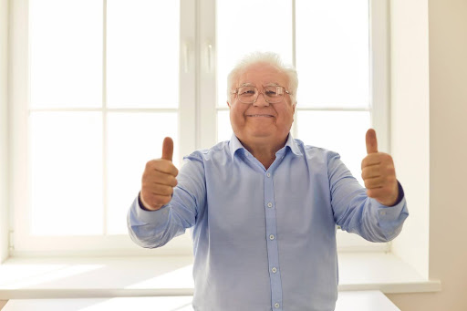 Smiling elderly man giving two thumbs up, standing in front of a sunlit window—symbolizing the challenges of identifying true wellness versus pretending to feel fine to avoid concern.