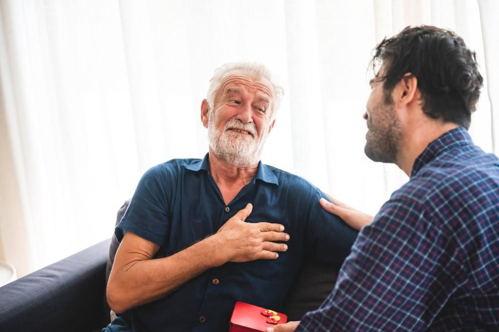 A senior man smiling with his hand on his chest as a younger man offers emotional support, showing warmth and connection in a caregiving moment.