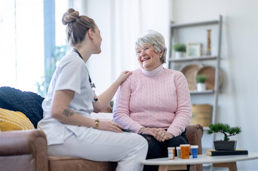 A cheerful elderly woman in a pink sweater talks with a caregiver at home, with a variety of pill bottles, including Tylenol, on the table in front of them—highlighting common over-the-counter habits among seniors.