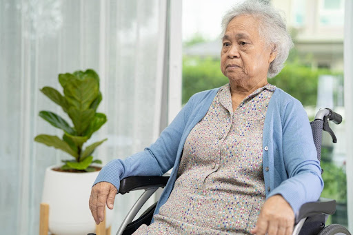 Elderly woman in a wheelchair sitting pensively in a bright room, reflecting the emotional challenges some seniors face around daily routines like taking a shower.