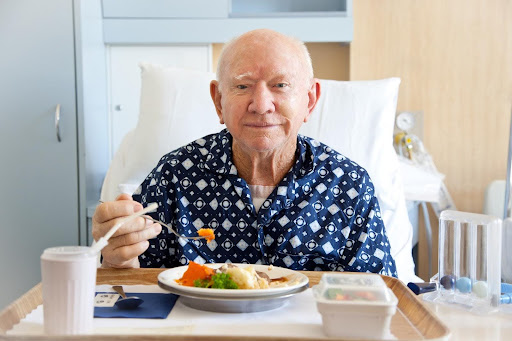 An elderly man in patterned pajamas sits upright in a hospital bed, smiling as he eats a meal from a tray. The image reflects compassionate care for seniors facing challenges like missing teeth, swallowing difficulties, or appetite loss—highlighting the importance of tailored meal support in elder care.
