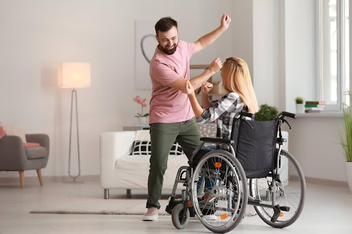A man joyfully dancing with a woman in a wheelchair inside a bright, cozy living room, illustrating the uplifting energy of seated Zumba and inclusive fitness for people with mobility challenges.