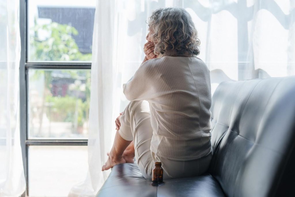 Lonely senior woman sitting by a window, looking out in deep thought with a medicine bottle nearby—capturing the emotional toll when family visits decline and the vital role of companionship.