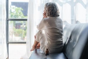 Lonely senior woman sitting by a window, looking out in deep thought with a medicine bottle nearby—capturing the emotional toll when family visits decline and the vital role of companionship.