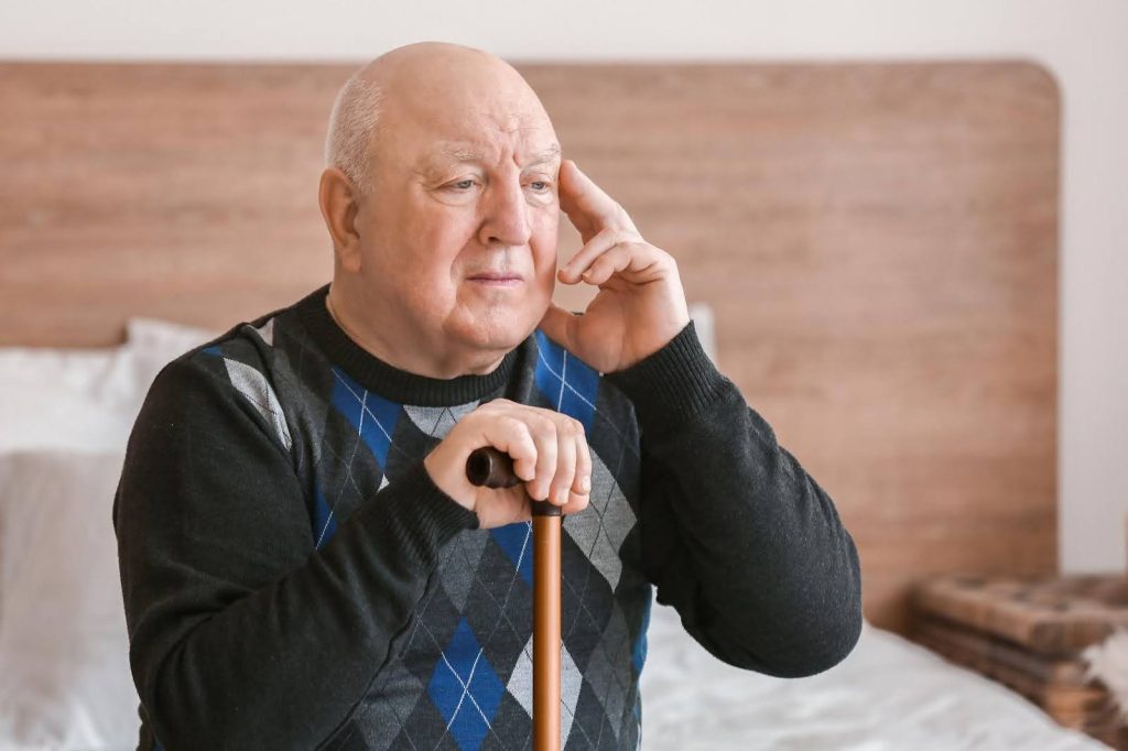 Elderly man sitting on a bed with a thoughtful expression, representing concern about whether Alzheimer’s disease is hereditary.