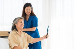 Filipino caregiver assisting a smiling elderly woman holding a mirror, symbolizing global trust in Filipino caregiving.
