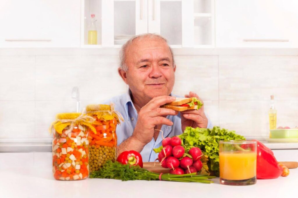 Smiling senior man enjoying a healthy sandwich surrounded by vegetables, juice, and preserved food jars, representing a customized diet for chronic illness.