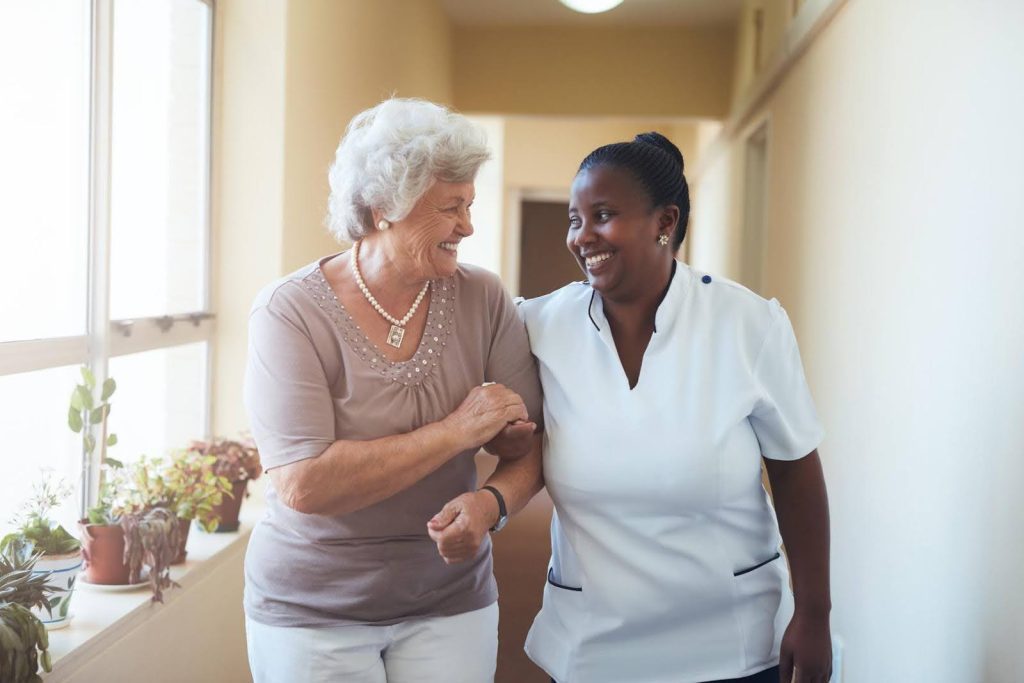 Caregiver walking arm-in-arm with a smiling senior woman, representing how friendship acts as medicine through the mental health benefits of companion care.