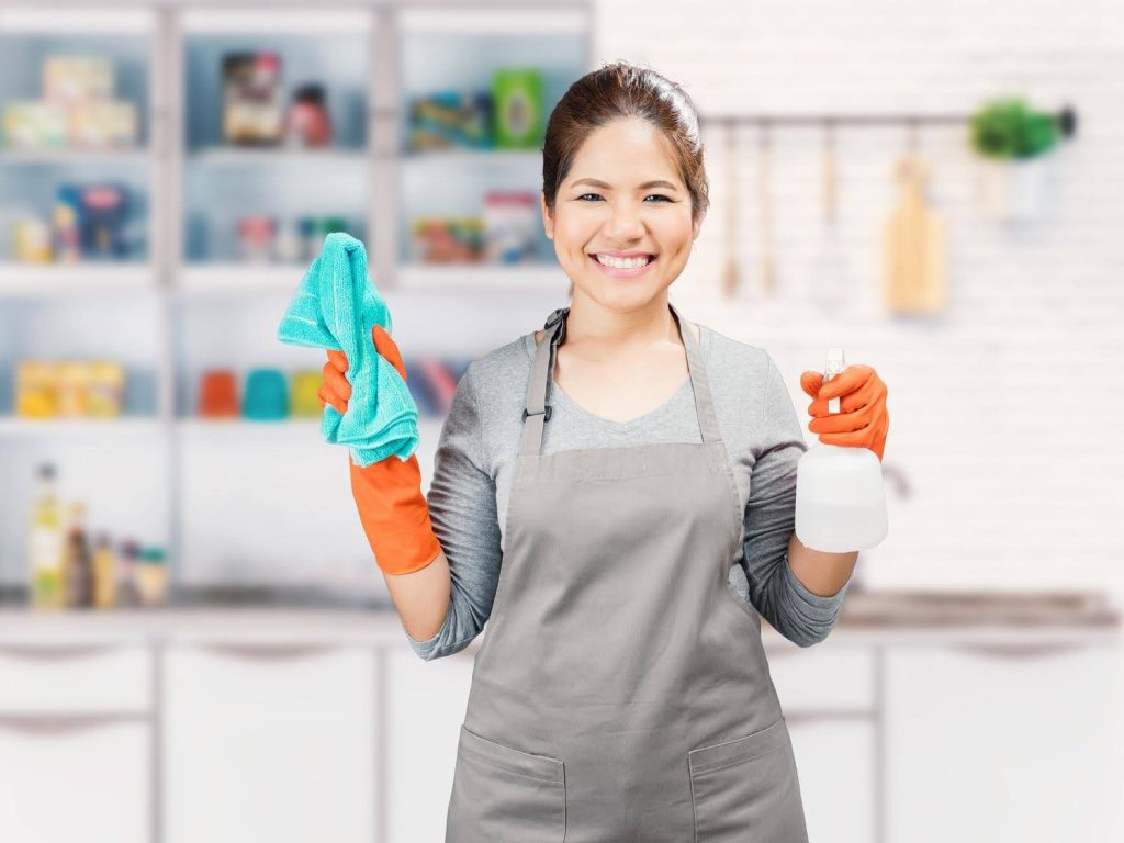 Smiling professional cleaner wearing gloves and apron, holding a spray bottle and cloth in a senior’s home kitchen—representing essential household support roles for aging adults.