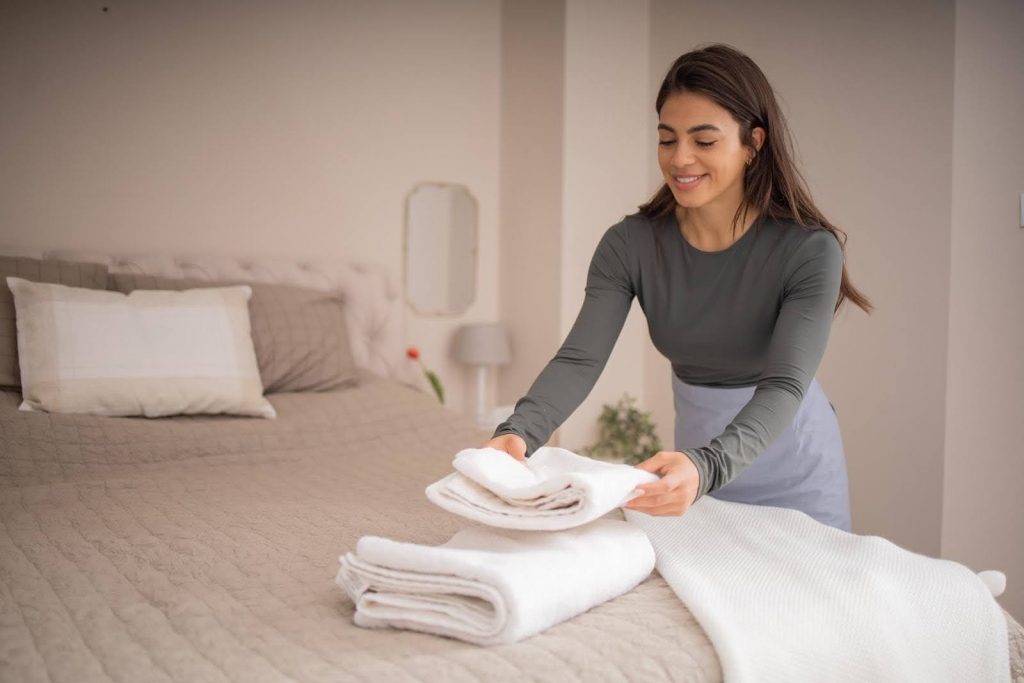 Smiling housekeeper folding towels in a neatly made bedroom, illustrating the importance of regular housekeeping help for seniors.
