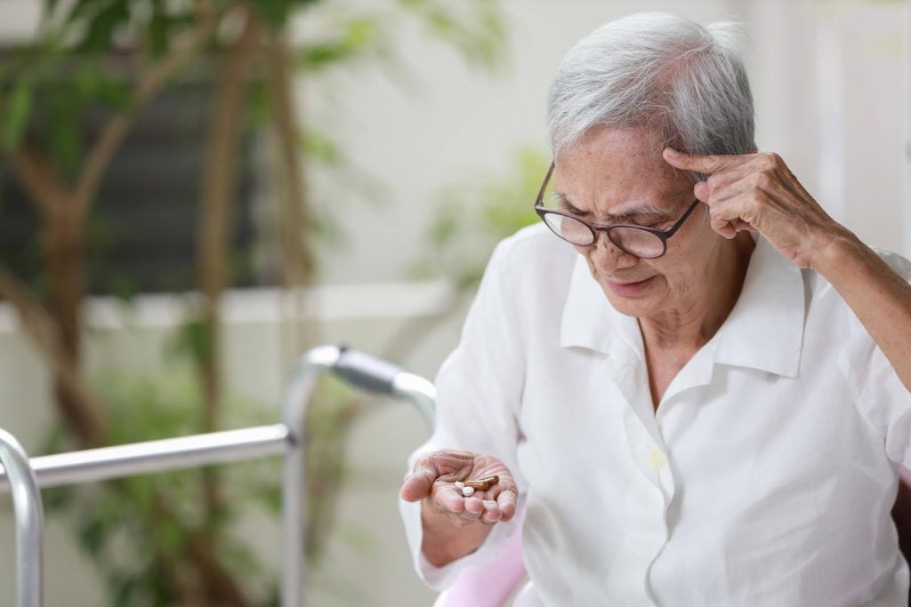 Elderly woman in glasses looking confused while holding meds in one hand and pointing to her temple—highlighting challenges with medication memory in seniors.