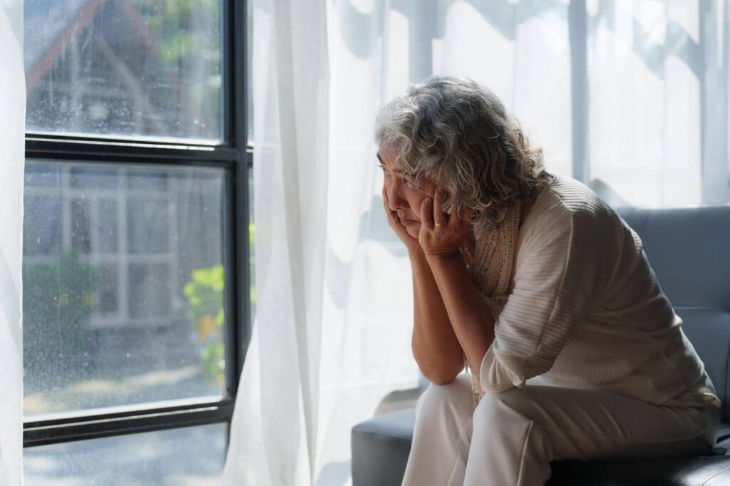 Elderly woman sitting alone and looking out a window, portraying the emotional impact of social deprivation among seniors.
