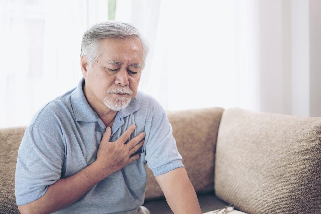 Elderly man sitting on a couch clutching his chest with a pained expression, illustrating the importance of timely health check-ins to detect critical conditions like strokes.