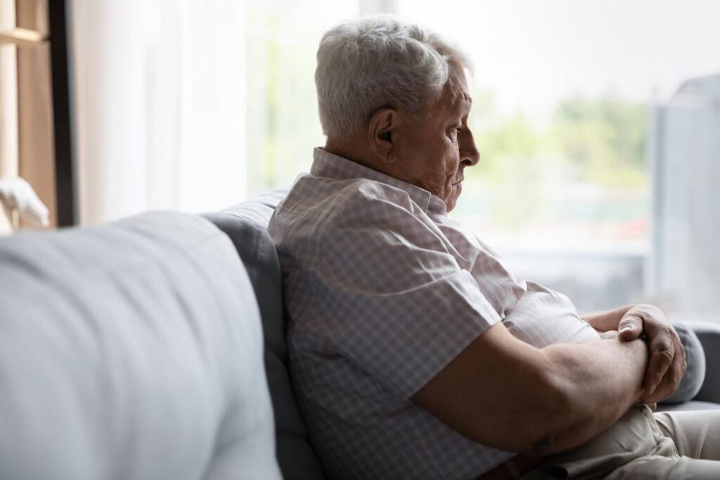 Sad senior man sitting alone on a couch with arms crossed, gazing downwards, illustrating possible signs of depress and loneliness in older adults.