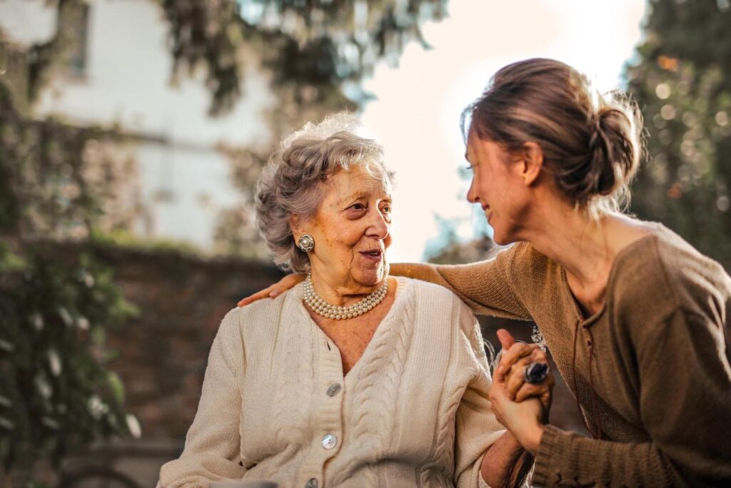 Adult daughter gently holding hands and talking with her elderly mother outdoors, symbolizing a caring conversation about personal care needs.