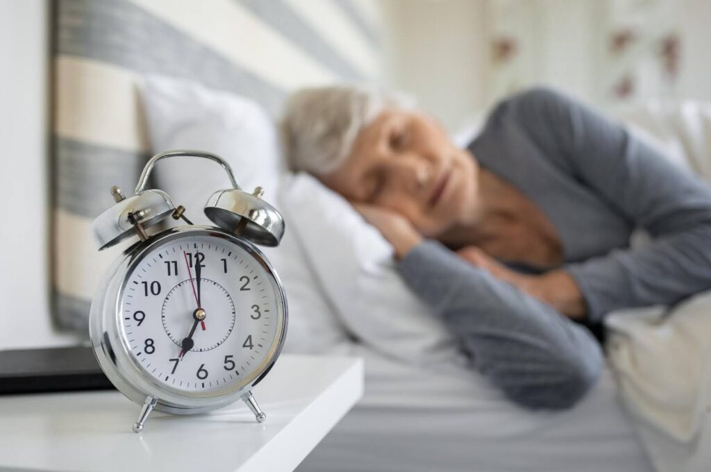 An elderly woman sleeps peacefully in bed while a classic alarm clock sits on the bedside table, illustrating concerns about excessive daytime sleep in seniors.