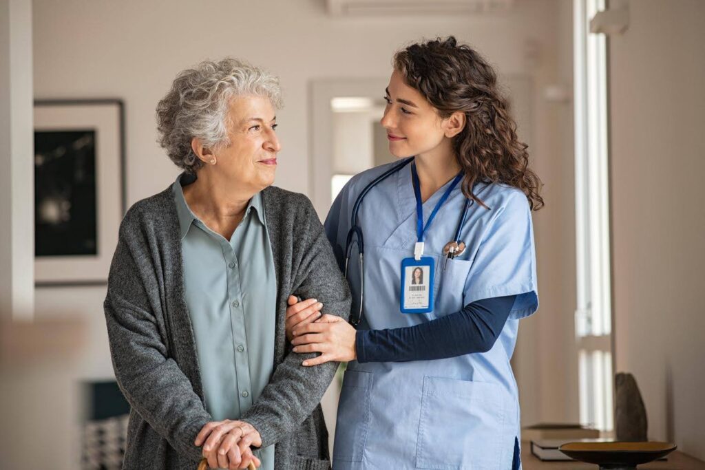 Smiling caregiver in blue scrubs supporting an elderly woman with a cane, representing weekend personal care options available when family caregivers need rest.