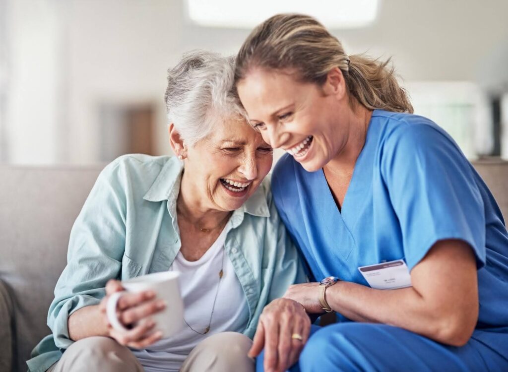 Smiling elderly woman sharing a joyful moment with her caregiver in blue scrubs, illustrating the importance of finding a good personality match for companion care.