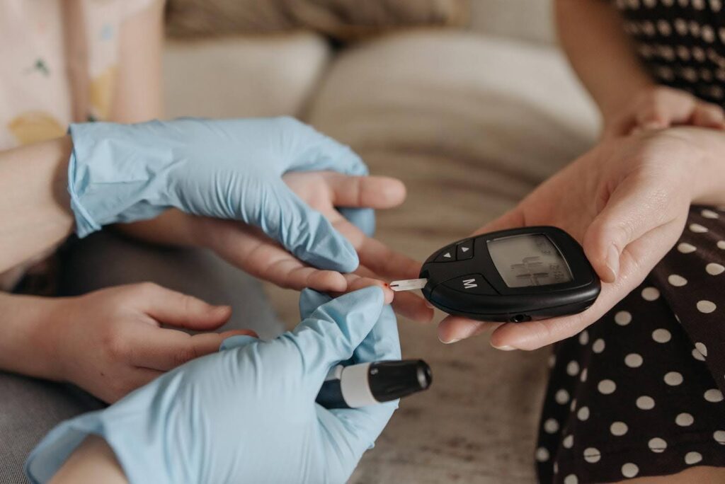 A caregiver wearing blue medical gloves checks a person’s blood sugar using a glucose meter, emphasizing the importance of daily home monitoring to prevent diabetic complications.