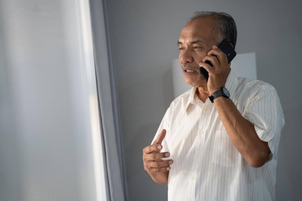 Senior man talking on the phone near a window, representing the importance of combining reassurance calls with emergency monitoring for safety and peace of mind.
