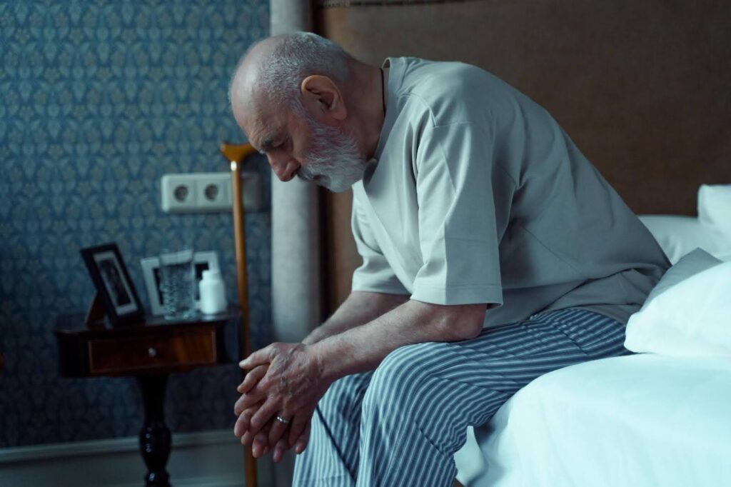 Elderly man sitting on the edge of his bed with head bowed and hands clasped, appearing deep in thought, symbolizing emotional struggles that may be hid from family and caregivers.