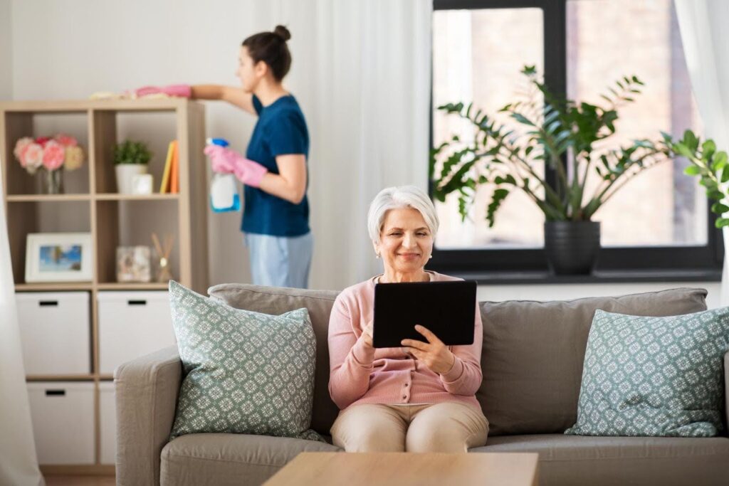 Elderly woman sitting on a couch using a tablet while a caregiver cleans in the background, illustrating clear boundaries in light housekeeping roles.