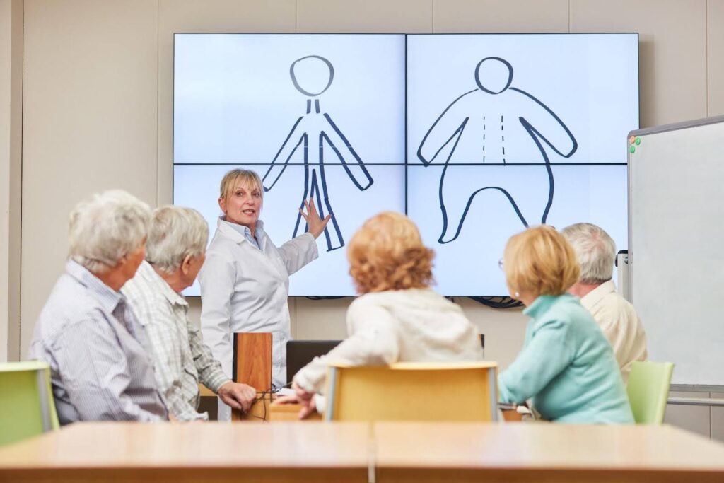 Healthcare professional presenting to a group of seniors with drawings of body outlines on a screen, illustrating hidden signs of malnutrition that are not always reflected on the scale.
