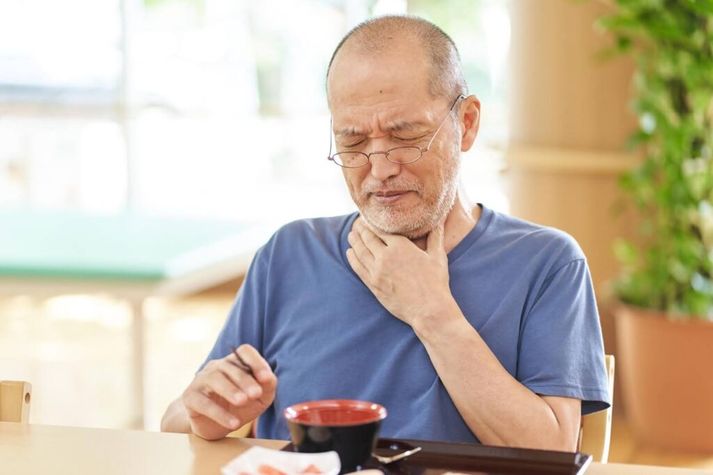 Elderly man in a blue shirt holding his throat with a pained expression while eating, illustrating challenges faced by those with dysphagia and the need for specialized meal preparation.