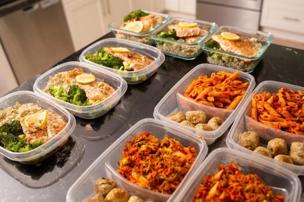 Multiple containers filled with neatly portioned meals including salmon with rice and broccoli, pasta with sauce, and vegetable mixes—showcasing organized meal prep as an act of care and nourishment.