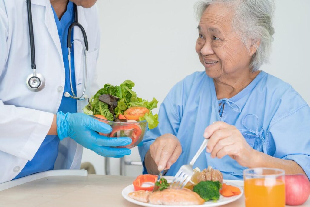 Smiling elderly woman in a hospital gown receiving a fresh salad from a healthcare professional, representing the need to address common nutrition myth concerns that may harm seniors' health.